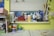 A cluttered workspace with various art and craft supplies arranged on a window sill. Items include containers of Mod Podge, paint, brushes, scissors, tape rolls, and a mug. The background has a window with closed blinds, and there's a yellow wall. Books and folders are stacked on the left side, and several paint cans and jars are on the table.