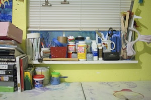 A cluttered workspace with various art and craft supplies arranged on a window sill. Items include containers of Mod Podge, paint, brushes, scissors, tape rolls, and a mug. The background has a window with closed blinds, and there's a yellow wall. Books and folders are stacked on the left side, and several paint cans and jars are on the table.