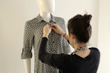 A person is adjusting the collar of a patterned shirt on a mannequin. The shirt has a black and white geometric design, and the mannequin is positioned against a neutral background.