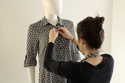 A person is adjusting the collar of a patterned shirt on a mannequin. The shirt has a black and white geometric design, and the mannequin is positioned against a neutral background.