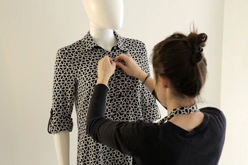 A person is adjusting the collar of a patterned shirt on a mannequin. The shirt has a black and white geometric design, and the mannequin is positioned against a neutral background.