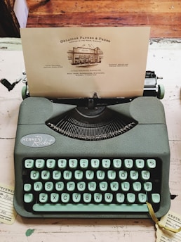 A vintage Hermes Rocket typewriter with mint-green keys sits on a desk. A piece of paper is inserted, displaying a print from Oblation Papers & Press, featuring details of its services and location. The background includes sections of a wooden floor and a rustic desk surface.