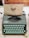 A vintage Hermes Rocket typewriter with mint-green keys sits on a desk. A piece of paper is inserted, displaying a print from Oblation Papers & Press, featuring details of its services and location. The background includes sections of a wooden floor and a rustic desk surface.