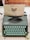 A vintage Hermes Rocket typewriter with mint-green keys sits on a desk. A piece of paper is inserted, displaying a print from Oblation Papers & Press, featuring details of its services and location. The background includes sections of a wooden floor and a rustic desk surface.