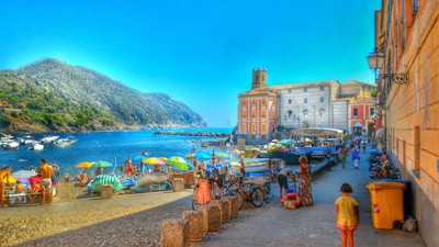 A vibrant beach scene with colorful umbrellas and people enjoying the sunny weather. The beach is lined with boats and a charming waterfront town with historic buildings. Hills rise in the background, adding a natural contrast to the bustling shoreline.