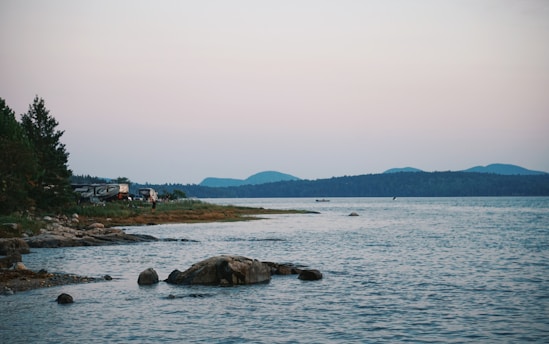 A tranquil lakeside scene with rocky shores and forested areas. A few camping trailers are visible among the trees near the water's edge, accompanied by a person standing on the shore. The background features distant mountains under a soft pastel-hued sky.