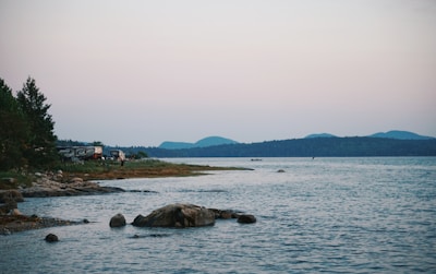 A tranquil lakeside scene with rocky shores and forested areas. A few camping trailers are visible among the trees near the water's edge, accompanied by a person standing on the shore. The background features distant mountains under a soft pastel-hued sky.