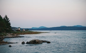 A tranquil lakeside scene with rocky shores and forested areas. A few camping trailers are visible among the trees near the water's edge, accompanied by a person standing on the shore. The background features distant mountains under a soft pastel-hued sky.