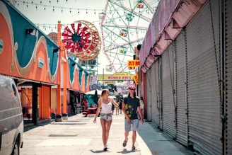 A lively amusement park scene with a Ferris wheel in the background. Two people walk along a pathway lined with bright, colorful arcade walls. The right side features closed shutters, and an ATM sign is visible.