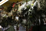 Plastic bags filled with various dried herbs hang in a market stall. Each bag contains different types and sizes of dried leaves, some labeled with small green stickers. The setting appears dimly lit, with shadows highlighting the textures of the herbs. Decorative items like ribbons and pine cones are interspersed among the hanging bags.