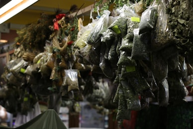 Plastic bags filled with various dried herbs hang in a market stall. Each bag contains different types and sizes of dried leaves, some labeled with small green stickers. The setting appears dimly lit, with shadows highlighting the textures of the herbs. Decorative items like ribbons and pine cones are interspersed among the hanging bags.