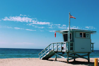 A lifeguard tower is situated on a sandy beach next to the ocean under a bright blue sky. The tower is painted in light blue and has an American flag waving at the top. The surrounding area is clear with a view of the horizon where the sea meets the sky.