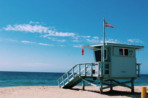 A lifeguard tower is situated on a sandy beach next to the ocean under a bright blue sky. The tower is painted in light blue and has an American flag waving at the top. The surrounding area is clear with a view of the horizon where the sea meets the sky.