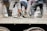 Several workers are engaged in a labor-intensive task on a dusty platform. They wear casual clothing, and their legs and footwear are visible, covered in dust and dirt. A thick rope is coiled on the ground, and a sense of manual labor and physical effort is evident.