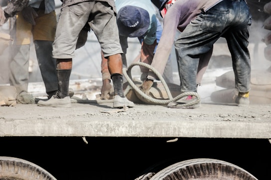 Several workers are engaged in a labor-intensive task on a dusty platform. They wear casual clothing, and their legs and footwear are visible, covered in dust and dirt. A thick rope is coiled on the ground, and a sense of manual labor and physical effort is evident.