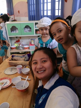 A group of people, including a smiling woman in a blue and white shirt, are in a room decorated with blue curtains. There are several children wearing costumes or uniforms, with some of them gathered around a table set with bowls, plates, and cups. The background shows kitchen utensils and shelves filled with items. The environment suggests a playful or educational setting.