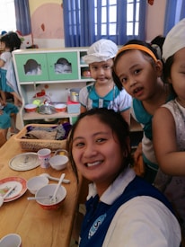 A group of people, including a smiling woman in a blue and white shirt, are in a room decorated with blue curtains. There are several children wearing costumes or uniforms, with some of them gathered around a table set with bowls, plates, and cups. The background shows kitchen utensils and shelves filled with items. The environment suggests a playful or educational setting.