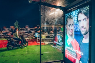 A busy street scene at night with a woman on a scooter, pedestrians moving swiftly in the background, and a brightly lit advertisement display on a bus stop. The ad features two people and is vividly illuminated against the darker surroundings. Reflections can be seen on the wet glass of the shelter, suggesting recent rain. The scene conveys a sense of urban hustle with people on the go.