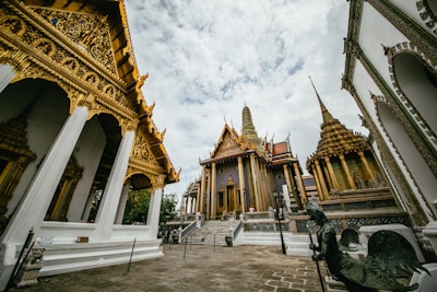 Intricate and ornate architecture characterized by rich golden details and pointed spires. The scene features multiple temple structures with lavish decorations under a partly cloudy sky. Visitors are observing the surroundings, enhancing the grandeur and scale of the location.
