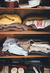 A wooden shelf holds neatly folded clothes, including sweaters with patterns, button-down shirts, and other garments. The top two shelves contain a mixture of casual and formal wear in various colors. The bottom shelf displays a few small containers and accessories.