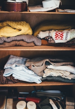 A wooden shelf holds neatly folded clothes, including sweaters with patterns, button-down shirts, and other garments. The top two shelves contain a mixture of casual and formal wear in various colors. The bottom shelf displays a few small containers and accessories.