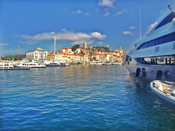 A picturesque harbor scene with numerous boats and yachts docked in the marina. The waterfront is lined with a variety of buildings, including residential and commercial structures, set against a backdrop of lush green hills and a clear blue sky with scattered white clouds. The water is a vivid blue, reflecting the sky's hues.