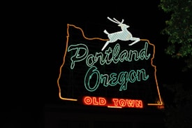 A neon sign with the words 'Portland Oregon' outlined in green, and 'Old Town' illuminated in red at the bottom. The outline of the state of Oregon is highlighted in orange and a white deer is depicted leaping at the top. The sign stands out against a dark night sky with a small portion of foliage visible on the right side.