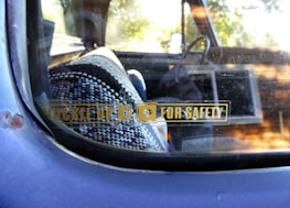 A close-up view of a car window with a safety reminder sticker that reads 'Buckle Up For Safety'. Inside the vehicle, a patterned woven seat cover is visible, along with a portion of the steering wheel. The window shows some wear and has a reflective surface.