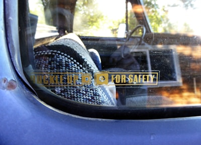 A close-up view of a car window with a safety reminder sticker that reads 'Buckle Up For Safety'. Inside the vehicle, a patterned woven seat cover is visible, along with a portion of the steering wheel. The window shows some wear and has a reflective surface.