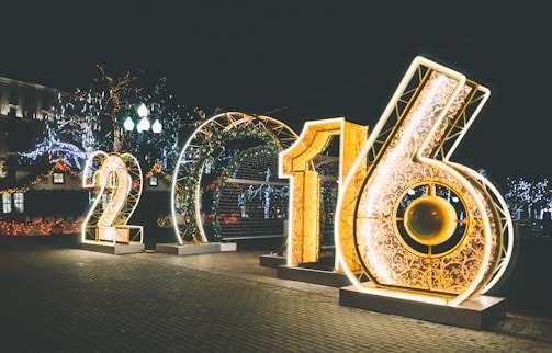 Large illuminated numbers '2016' are displayed in an outdoor setting at night, surrounded by decorative string lights and festive decorations in the background. The numbers are ornately designed with intricate patterns and emit a warm glow.