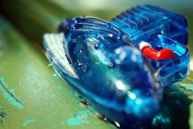 A close-up view of a blue plastic toy resembling a futuristic vehicle is placed on a green surface. Small red and white components are visible inside the transparent body of the toy. Water droplets are scattered across the surface, adding a glossy and fresh appearance.