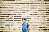 A young person is standing in front of a large, yellow apartment building. The structure has many windows and air conditioning units. The individual is wearing a blue shirt and a backpack, looking to the side with an urban backdrop.
