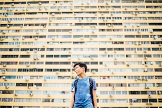 A young person is standing in front of a large, yellow apartment building. The structure has many windows and air conditioning units. The individual is wearing a blue shirt and a backpack, looking to the side with an urban backdrop.