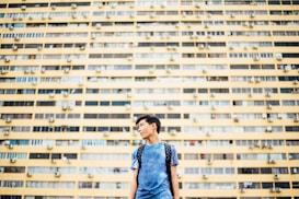 A young person is standing in front of a large, yellow apartment building. The structure has many windows and air conditioning units. The individual is wearing a blue shirt and a backpack, looking to the side with an urban backdrop.