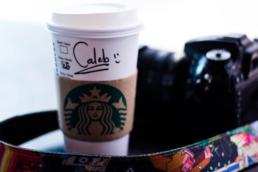 A paper coffee cup with a sleeve and a handwritten name sits next to a camera with a colorful strap. The sleeve displays a prominent green logo of a coffee brand.