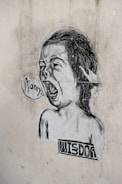 A black and white street art illustration featuring a child-like face with an open mouth, as if shouting. The word 'money' is written in a speech bubble. Below the face, the word 'wisdom' is printed in a blocky font. The image is pasted on a textured, worn wall.
