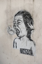 A black and white street art illustration featuring a child-like face with an open mouth, as if shouting. The word 'money' is written in a speech bubble. Below the face, the word 'wisdom' is printed in a blocky font. The image is pasted on a textured, worn wall.