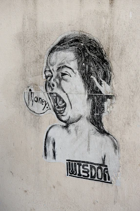 A black and white street art illustration featuring a child-like face with an open mouth, as if shouting. The word 'money' is written in a speech bubble. Below the face, the word 'wisdom' is printed in a blocky font. The image is pasted on a textured, worn wall.