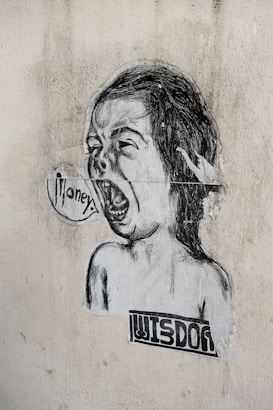 A black and white street art illustration featuring a child-like face with an open mouth, as if shouting. The word 'money' is written in a speech bubble. Below the face, the word 'wisdom' is printed in a blocky font. The image is pasted on a textured, worn wall.