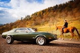 A vintage car is parked on a rustic road, set against a backdrop of rolling hills with autumn-colored trees. A person rides a horse along the road, creating a juxtaposition of classic automotive and equestrian elements.