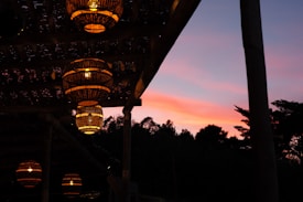 Several woven lanterns are hanging from a wooden pergola, casting a warm light. The evening sky in the background displays hues of pink and purple, creating a striking contrast with the dark silhouettes of nearby trees and foliage.