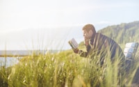 man reading book on beach near lake during daytime