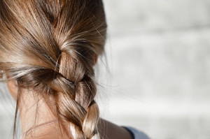 A close-up view of a person's hair styled in a neat braid. The hair appears light brown with hints of blonde highlights, and the texture is smooth and shiny. The background is a soft, out-of-focus grey, which contrasts with the sharp detail of the hair.