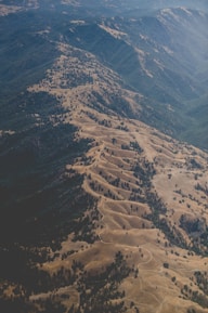 An aerial view of a mountainous and hilly landscape characterized by undulating ridges, valleys, and sparse tree coverage. The terrain appears dry, with shades of brown and green indicating a mix of vegetation and exposed earth.