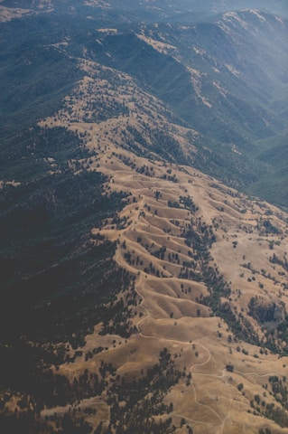 An aerial view of a mountainous and hilly landscape characterized by undulating ridges, valleys, and sparse tree coverage. The terrain appears dry, with shades of brown and green indicating a mix of vegetation and exposed earth.