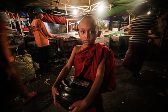 A young child wearing a red robe stands in a dimly lit market environment, holding a black container. The market is bustling with several people moving around, and there are stalls with food in the background. Overhead tarps and lights create a warm atmosphere with shadows and contrasting light.