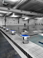 A spacious indoor swimming pool area with multiple lanes marked by black and white flags. Diving platforms are numbered and positioned at the pool's edge. The space is industrial with exposed piping and large lighting above. The walls have abstract line patterns and the overall setting is tidy and quiet.