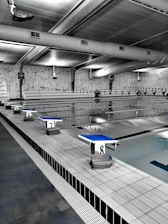 A spacious indoor swimming pool area with multiple lanes marked by black and white flags. Diving platforms are numbered and positioned at the pool's edge. The space is industrial with exposed piping and large lighting above. The walls have abstract line patterns and the overall setting is tidy and quiet.