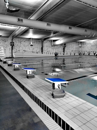 A spacious indoor swimming pool area with multiple lanes marked by black and white flags. Diving platforms are numbered and positioned at the pool's edge. The space is industrial with exposed piping and large lighting above. The walls have abstract line patterns and the overall setting is tidy and quiet.