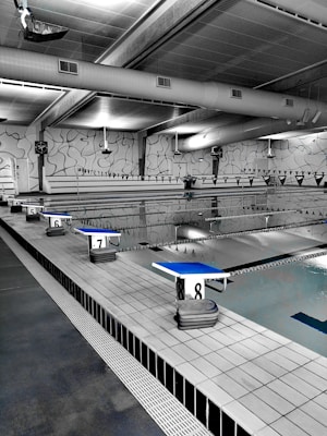 A spacious indoor swimming pool area with multiple lanes marked by black and white flags. Diving platforms are numbered and positioned at the pool's edge. The space is industrial with exposed piping and large lighting above. The walls have abstract line patterns and the overall setting is tidy and quiet.